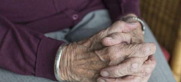 Close-up of elderly hands folded in the lap, wearing a purple cardigan and silver bracelets.