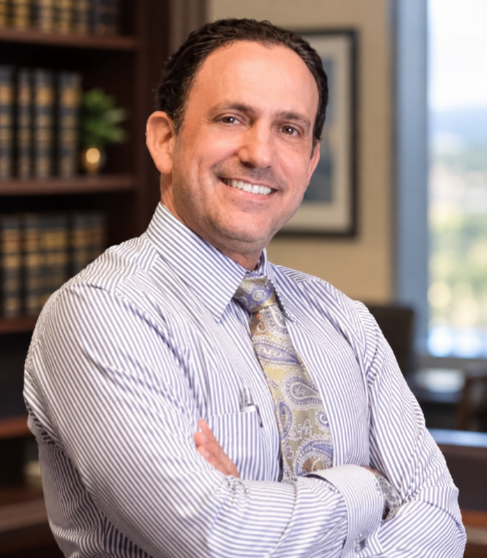 Smiling man in a striped dress shirt and paisley tie, arms crossed in an office setting.
